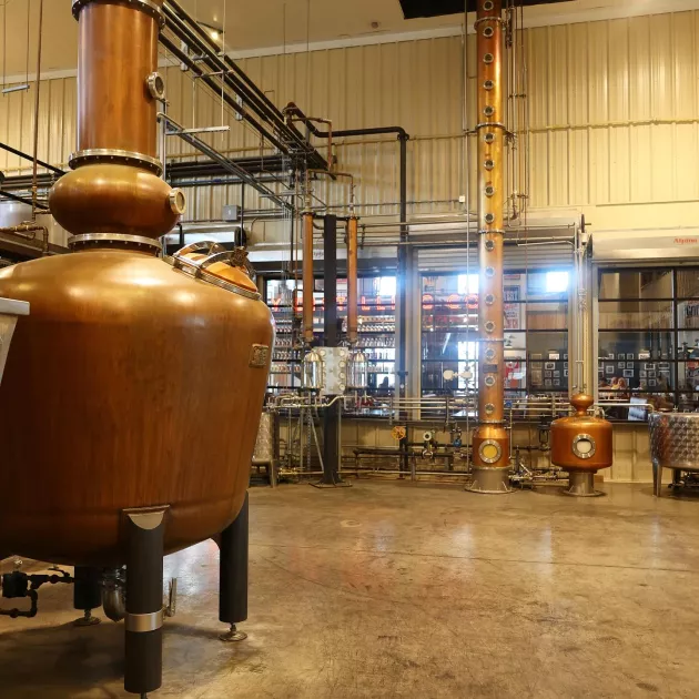 A copper column still inside Old Glory Distilling Co., showcasing the intricate distillation equipment used in crafting their spirits. The large still features multiple circular sight glasses, pipes, and gauges, with a view of the distillery's tasting room through glass windows in the background.