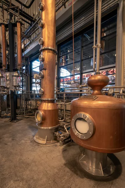 A close-up view of the copper column stills inside Old Glory Distilling Co., where whiskey is carefully distilled. The stills feature multiple sight glasses and polished copper details, with the distillery's neon-lit sign visible through the large windows in the background.